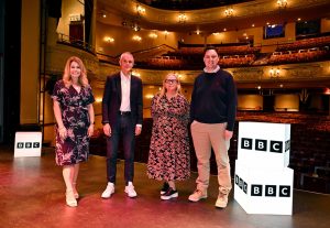 Left to Right: Blonde lady wearing a dress, white man wearing a suit, blonde lady with glasses wearing a dress and a man with dark hair wearing jumper and chinos on a stage with BBC lightboxes