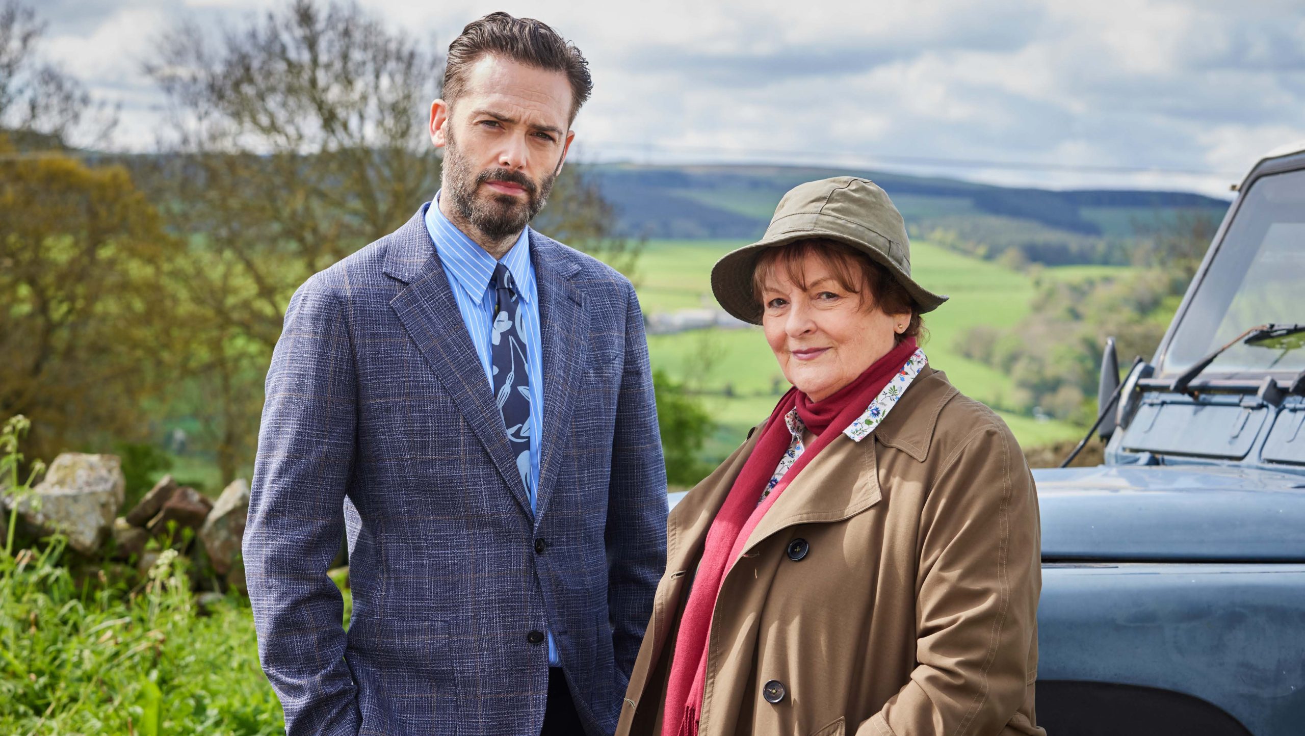 man with beard in a suit and older lady with trench coat stood in front of car in the countryside